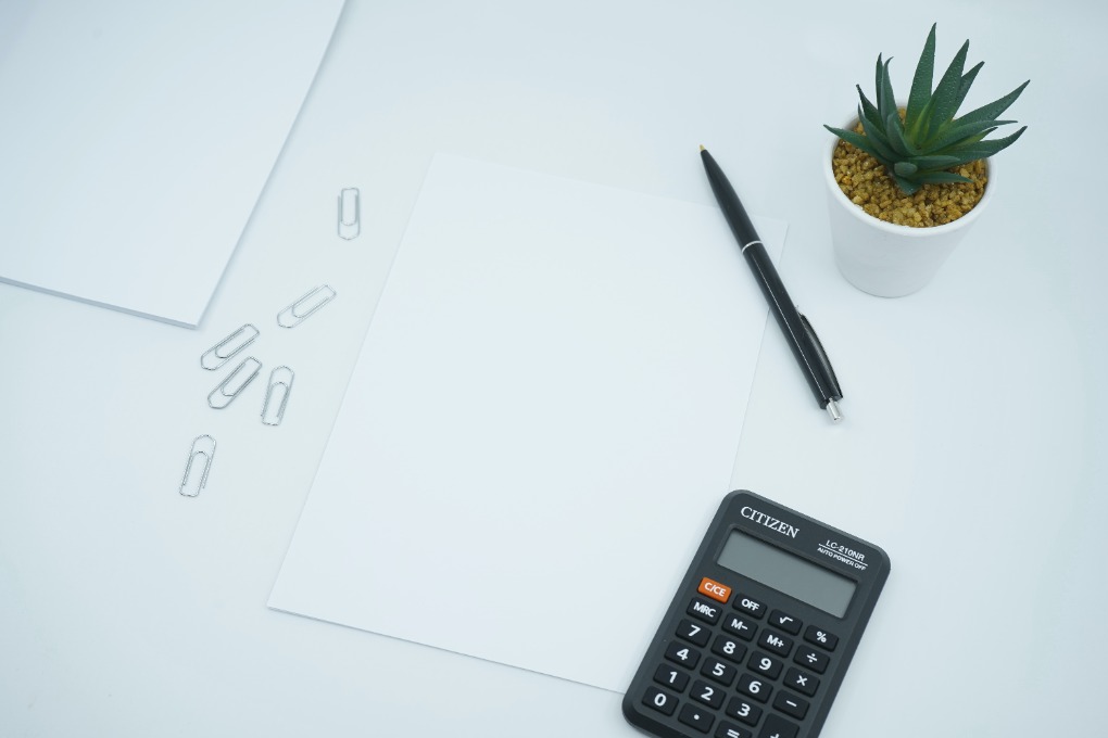 A picture of a black wooden table with a black pocket calculator, 2 white sheets of paper and a small white cactus pot
