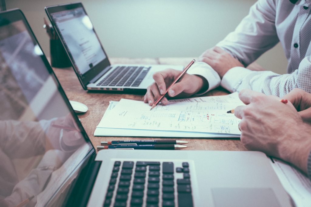 A picture of 2 people at a table with 2 computers and a picture of accounting graphs on them, you can only see hands, one hand with a pencil towards a page with some text