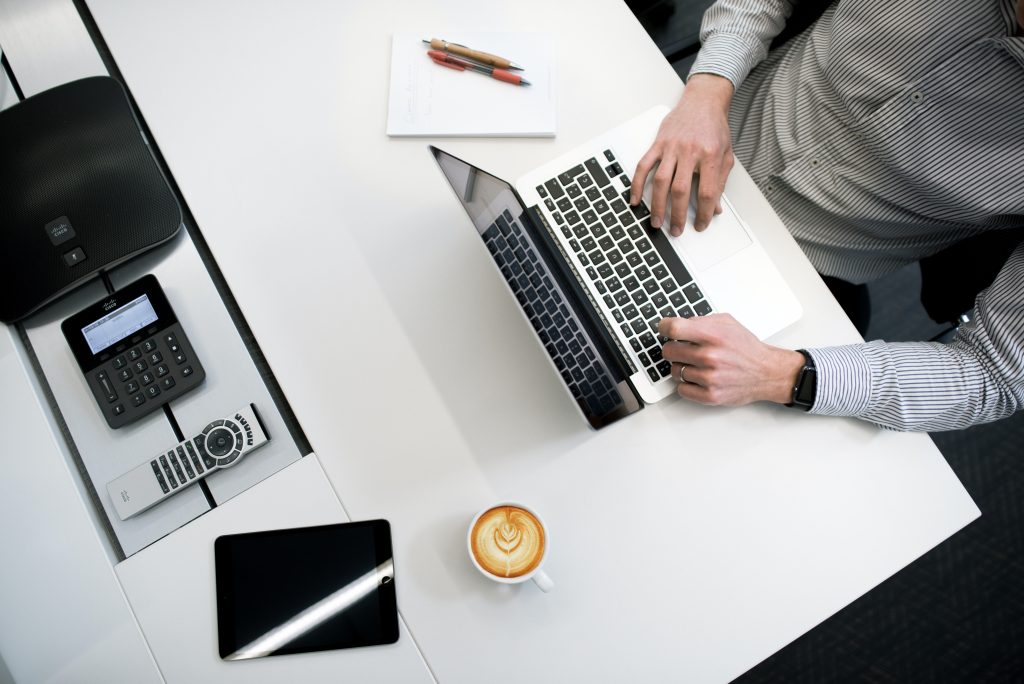 Overhead shot of a man sitting and typing on a computer on a white table with a computer and a cup of coffee
